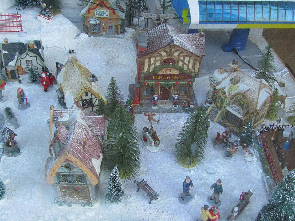 Miniature snowy Christmas village display, small decorated buildings, snow-covered rooftops, pine trees, tiny figurines of people, festive holiday scene, winter-themed setup