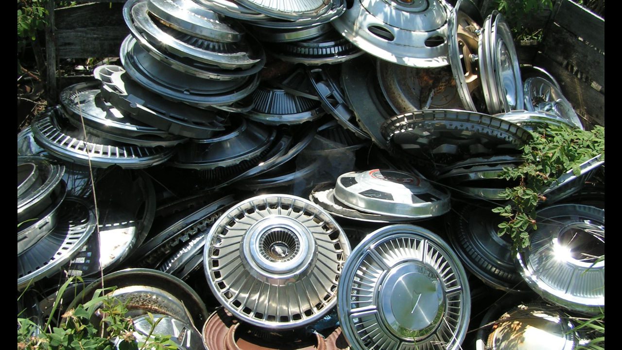 Pile of shiny, vintage metal hubcaps stacked haphazardly in an overgrown outdoor area, reflecting sunlight