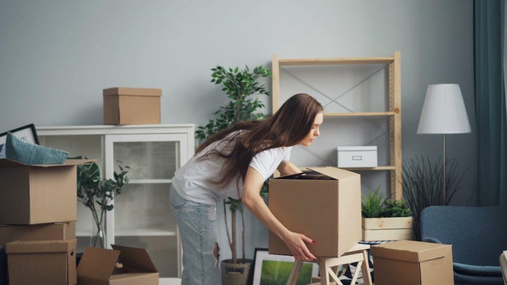 Woman bending over to lift a cardboard box, surrounded by several other boxes, in a living room with scattered items, empty shelves, and indoor plants, suggesting a space in transition or mid-organization