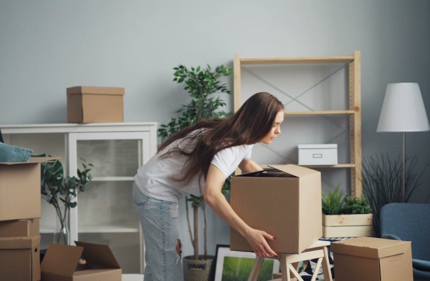 Woman bending over to lift a cardboard box, surrounded by several other boxes, in a living room with scattered items, empty shelves, and indoor plants, suggesting a space in transition or mid-organization