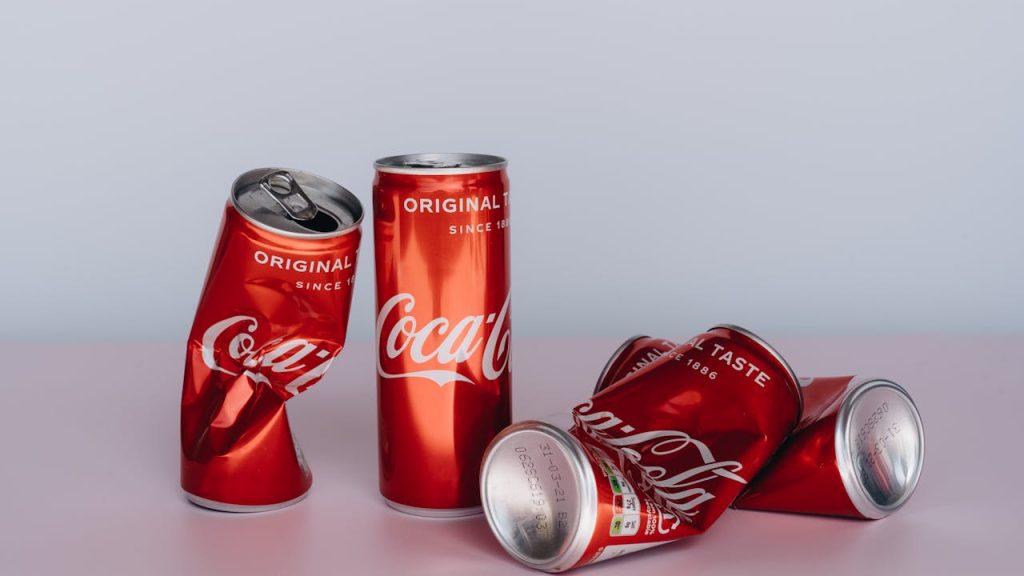 five Coca-Cola cans on a pink surface with a light gray background, two upright and three crushed and lying down