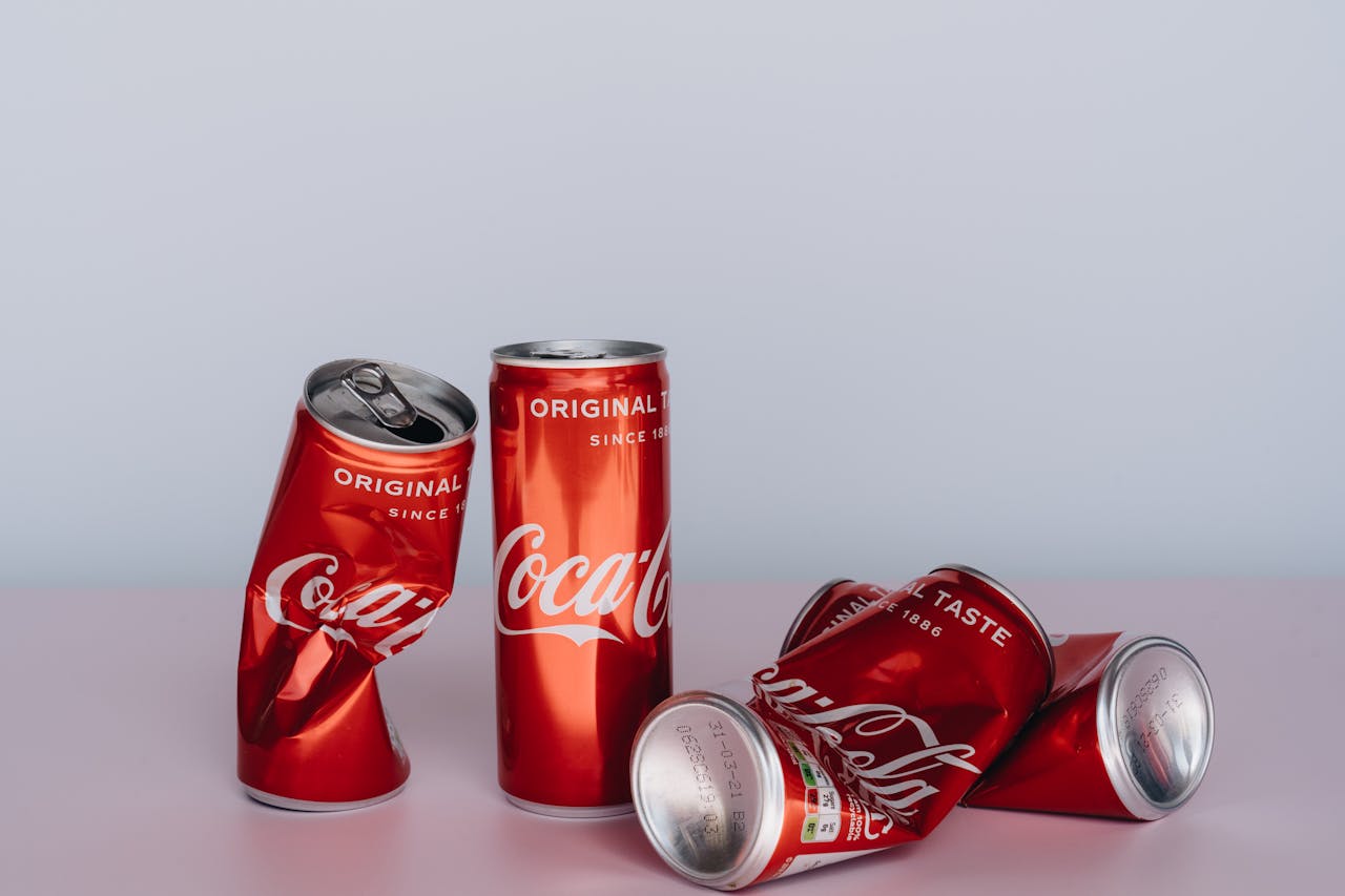 five Coca-Cola cans on a pink surface with a light gray background, two upright and three crushed and lying down