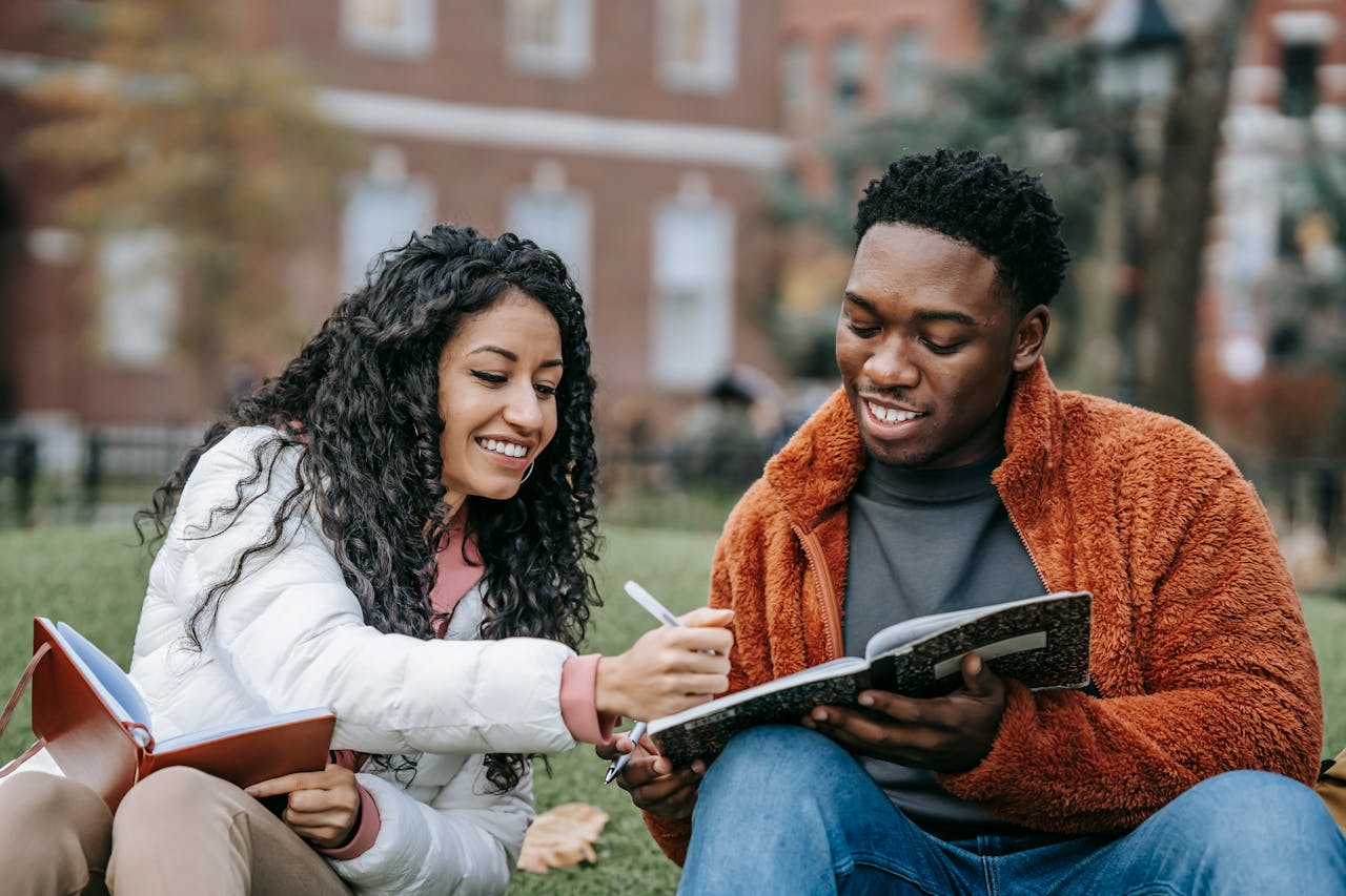 two college students sitting on grass, smiling and writing in notebooks while studying together outdoors