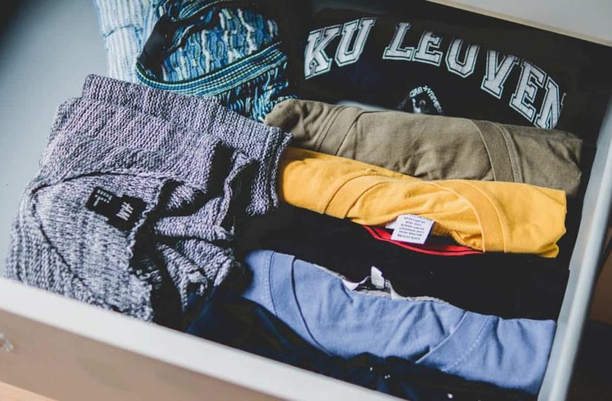 Open drawer filled with neatly folded T-shirts of various colors, including grey, yellow, blue, and black, with one shirt displaying "KU LEUVEN" text