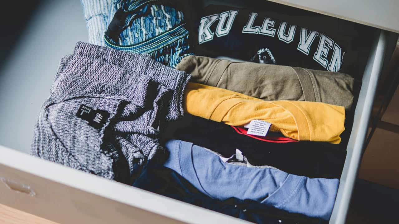 Open drawer filled with neatly folded T-shirts of various colors, including grey, yellow, blue, and black, with one shirt displaying "KU LEUVEN" text
