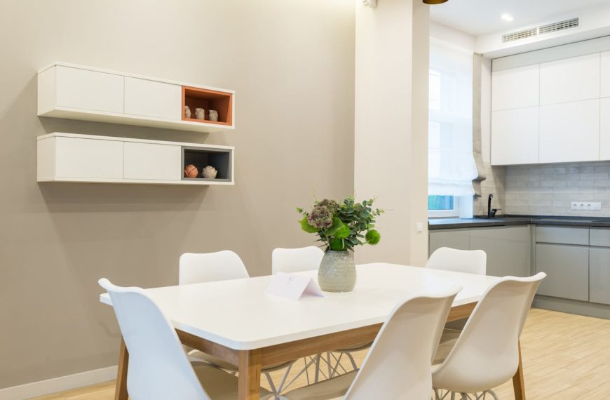 Modern dining area, white table with six white chairs, minimalist wall shelves, beige accent wall, vase with green and white flowers as centerpiece, bright natural lighting