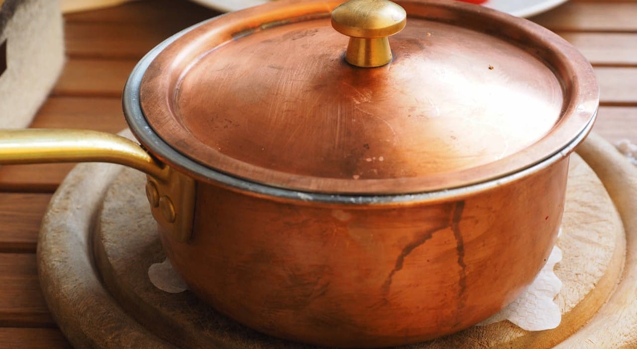 Copper saucepan with a lid, brass handle and knob, resting on a round wooden trivet, placed on a wooden table, blurred plate of food with tomato