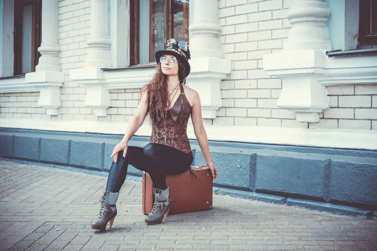 Woman in steampunk attire, wearing a brown corset vest, black leggings, heeled boots, and a decorated top hat with goggles, sitting on a vintage suitcase in front of a white brick building