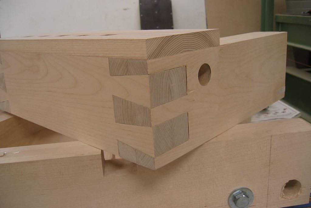 Close-up of dovetail joints on light wood panels, showing precise interlocking craftsmanship with a circular hole drilled into one of the panels