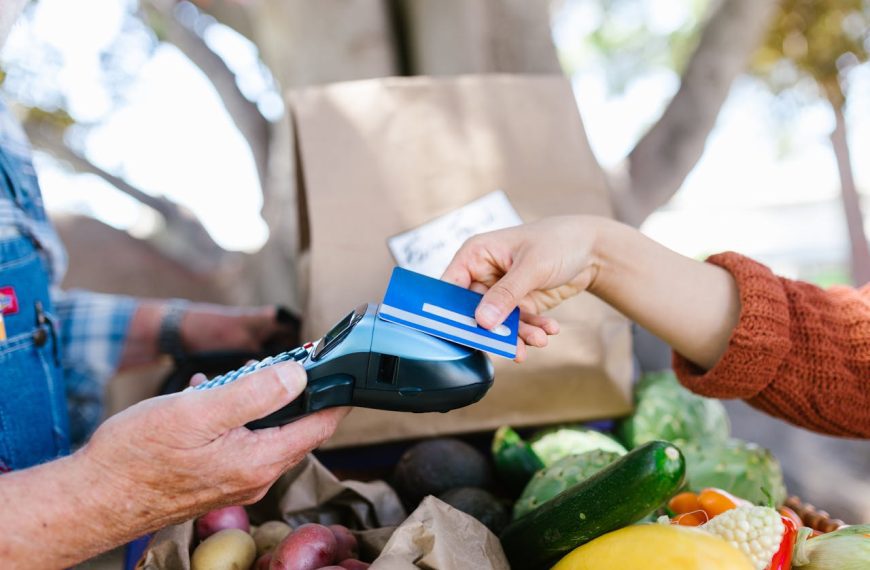 A person handing a blue credit card to a vendor using a card reader, fresh fruits and vegetables in the foreground, cardboard box and trees in the background