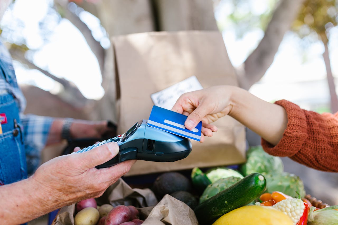 A person handing a blue credit card to a vendor using a card reader, fresh fruits and vegetables in the foreground, cardboard box and trees in the background
