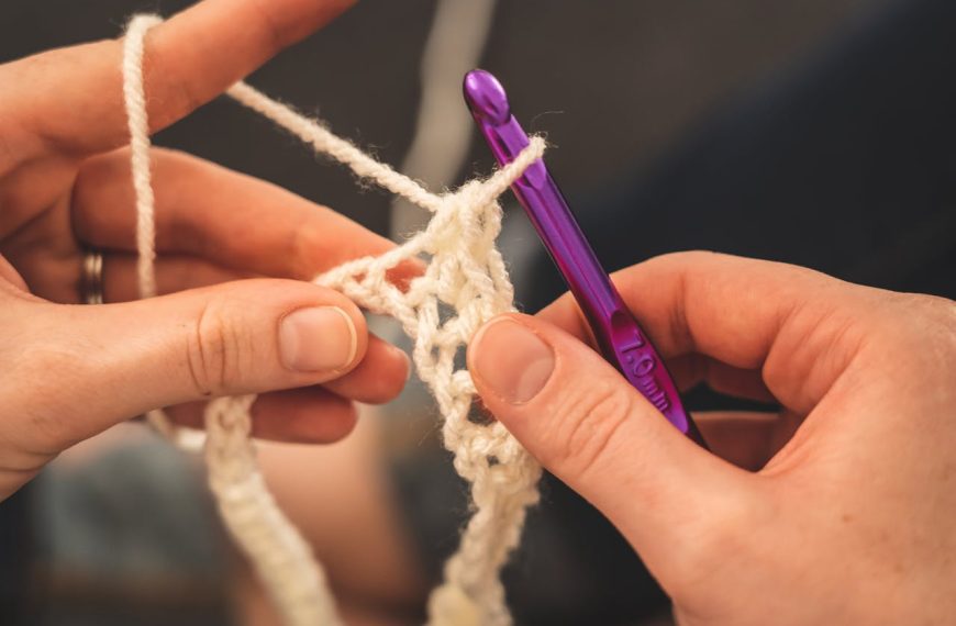 Close-up of hands crocheting with white yarn and a purple 7.0mm crochet hook, forming a blanket pattern