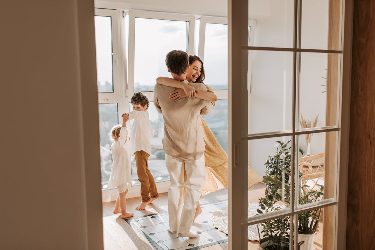 Family enjoying a joyful moment at home, with parents dancing in a loving embrace while two children play nearby on a patterned tile floor