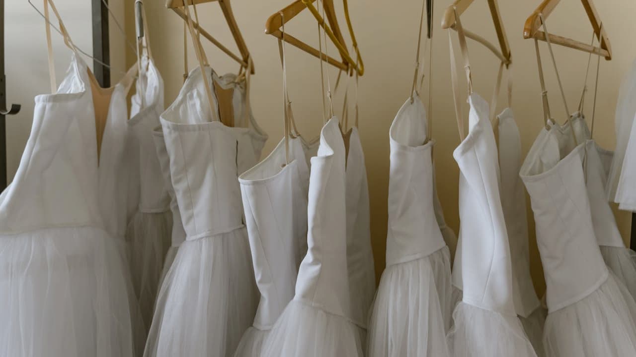 Row of white ballet costumes with tulle skirts hanging on wooden hangers against a beige wall