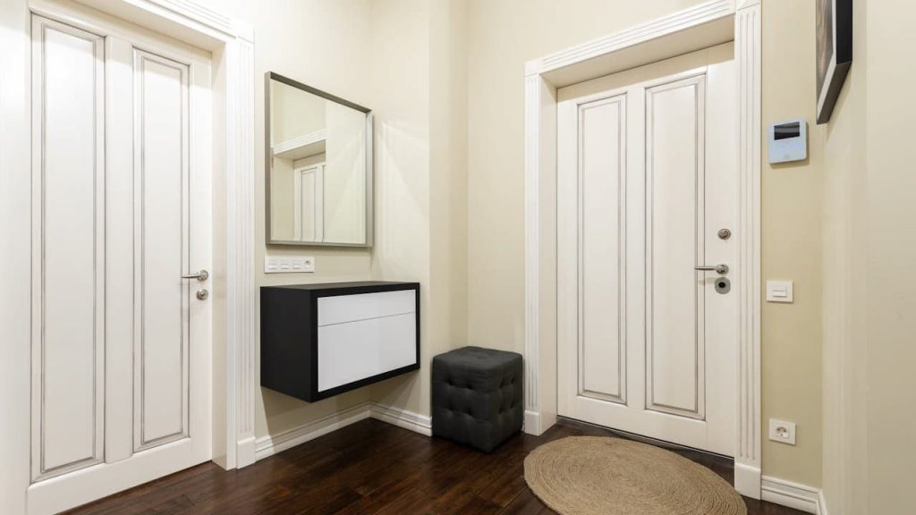 Modern entryway with cream-colored walls, two white double doors, dark wood flooring, black and white floating console, square mirror above console, black cushioned ottoman