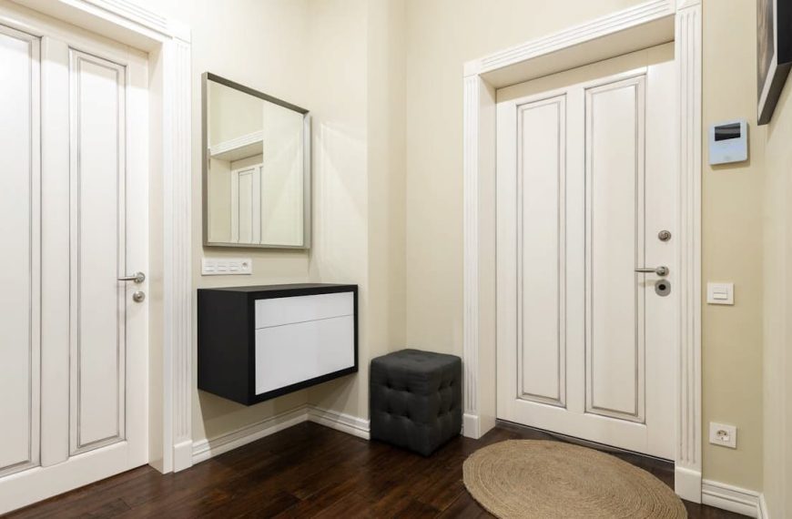 Modern entryway with cream-colored walls, two white double doors, dark wood flooring, black and white floating console, square mirror above console, black cushioned ottoman