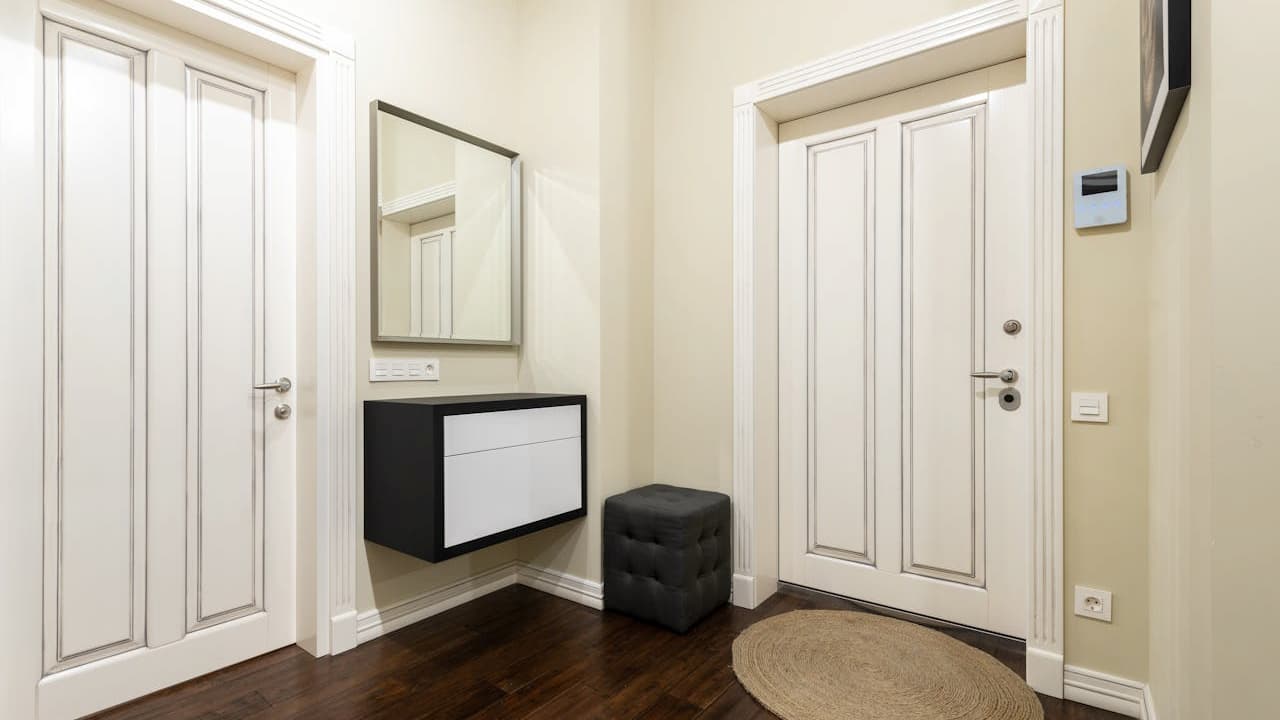 Modern entryway with cream-colored walls, two white double doors, dark wood flooring, black and white floating console, square mirror above console, black cushioned ottoman