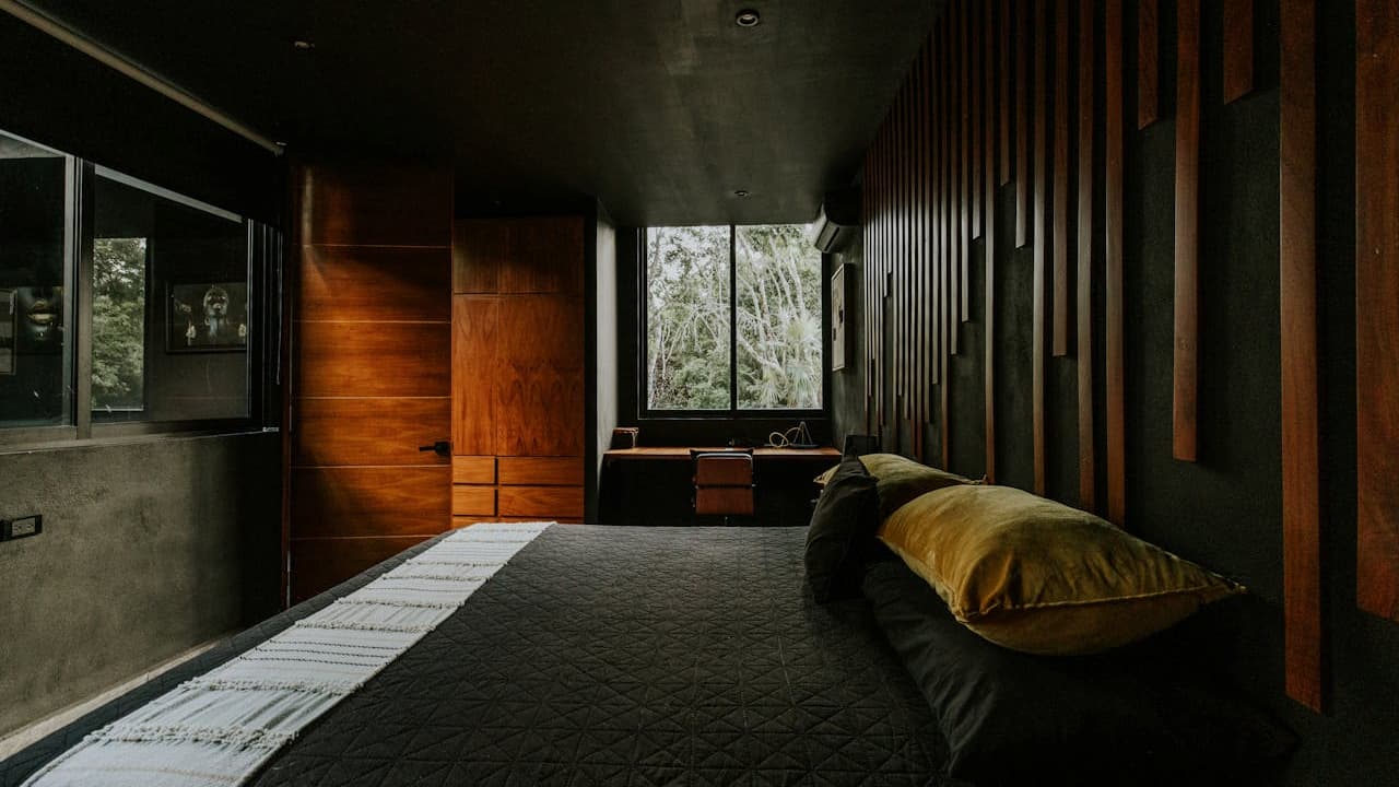 Modern bedroom with dark walls and ceiling, wood accent wall, large bed with black quilt and mustard pillow, wooden furniture, window with view of trees