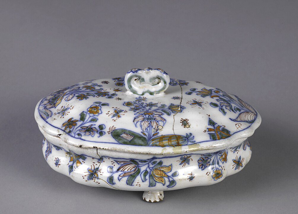 Decorative porcelain trinket box, oval-shaped, floral blue and orange pattern, ornate knob on lid, ceramic feet, placed on plain background