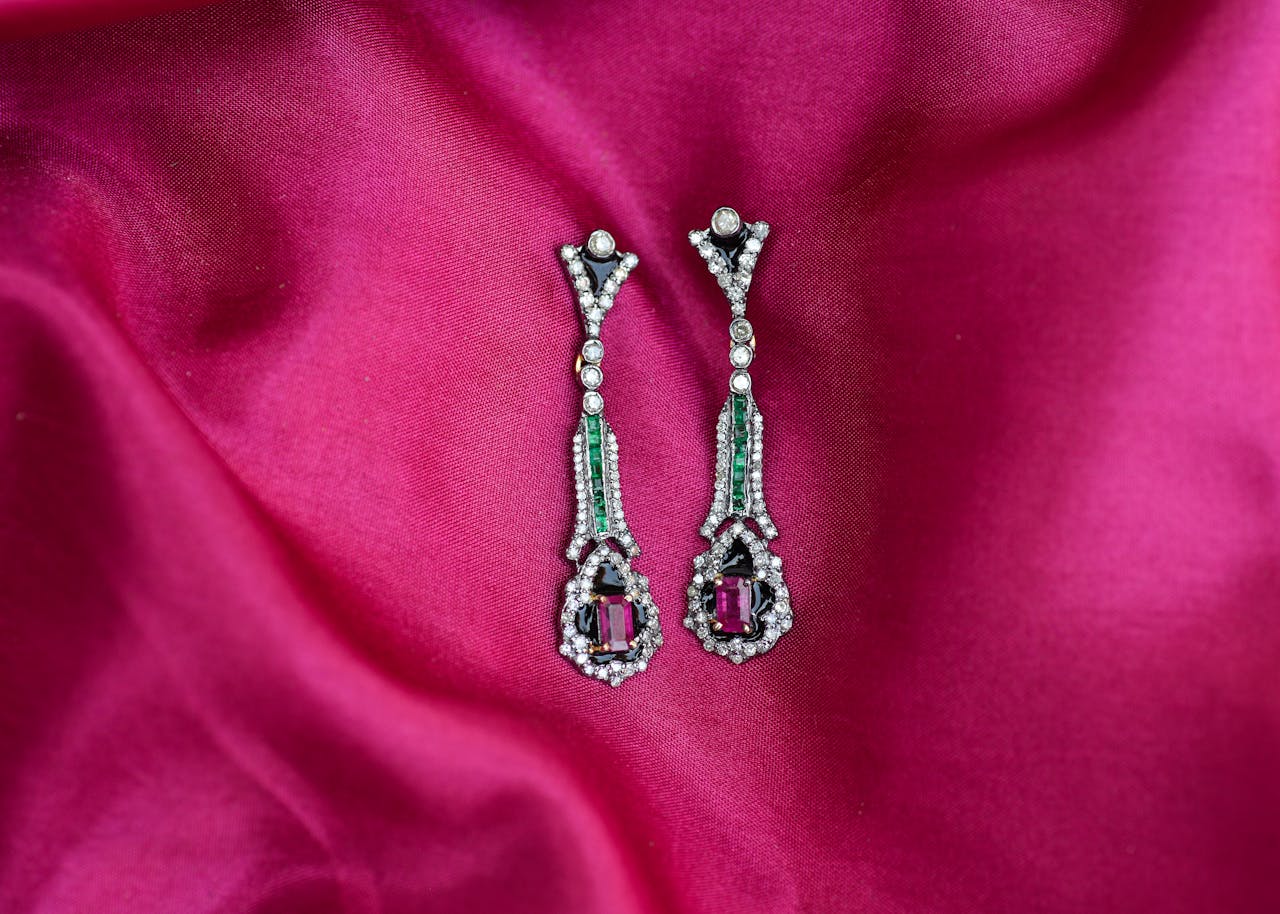 Elegant diamond and emerald drop earrings with deep pink gemstones, set against a rich, vibrant magenta silk fab