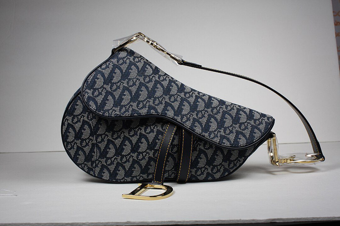 Designer handbag, dark blue patterned fabric, gold hardware accents, black leather strap, structured saddle shape, high-end fashion accessory
