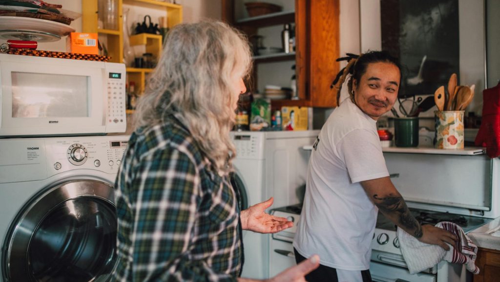 Two people are in a kitchen, one with long gray hair and a plaid shirt facing a man in a white shirt smiling while washing dishes, shelves with food items and kitchen appliances like a microwave and washing machine are visible, the mood appears casual and cheerful, possibly during a friendly conversation or shared chore time
