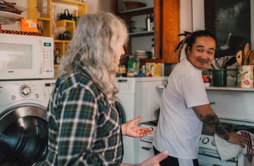 Two people are in a kitchen, one with long gray hair and a plaid shirt facing a man in a white shirt smiling while washing dishes, shelves with food items and kitchen appliances like a microwave and washing machine are visible, the mood appears casual and cheerful, possibly during a friendly conversation or shared chore time