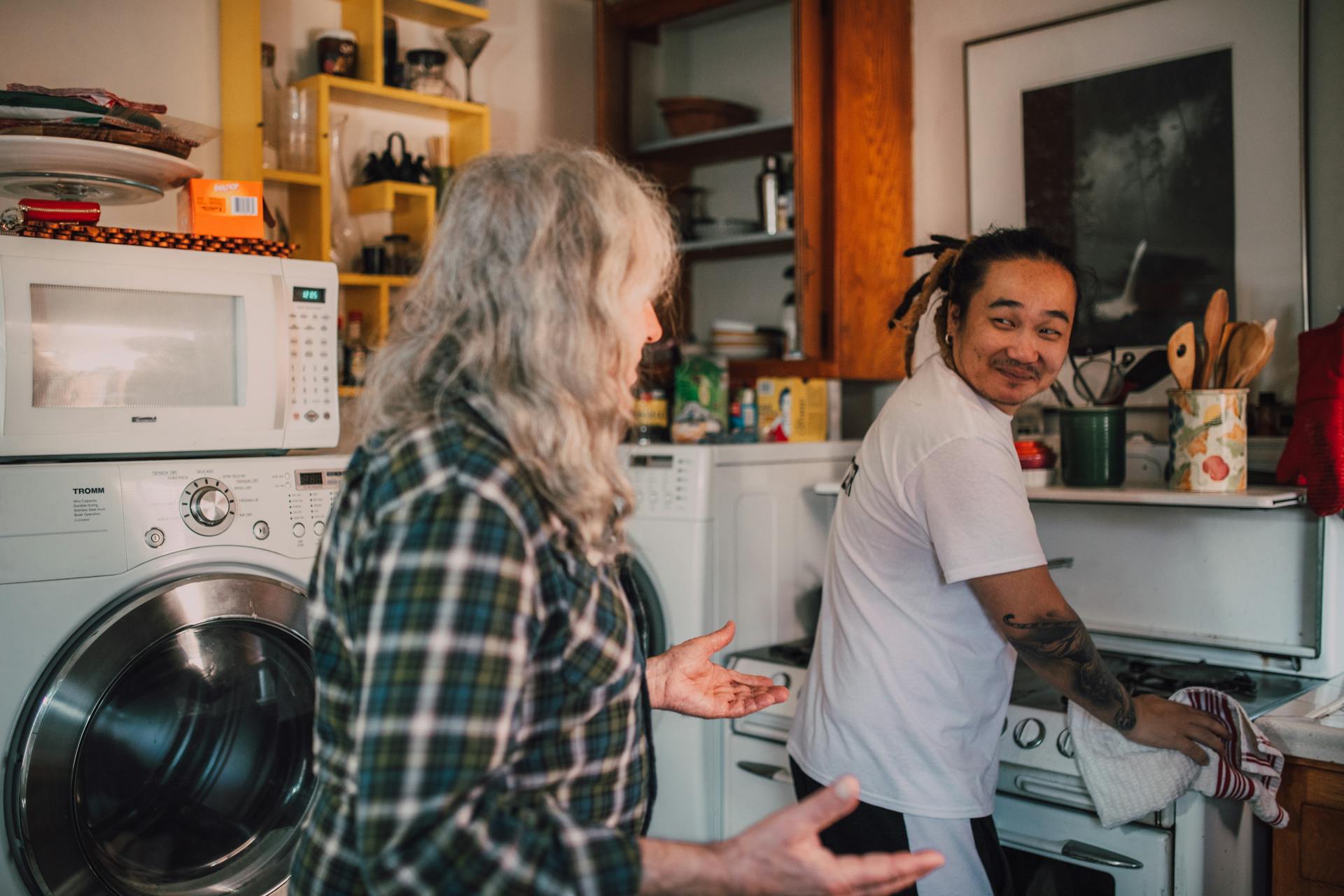 Two people are in a kitchen, one with long gray hair and a plaid shirt facing a man in a white shirt smiling while washing dishes, shelves with food items and kitchen appliances like a microwave and washing machine are visible, the mood appears casual and cheerful, possibly during a friendly conversation or shared chore time