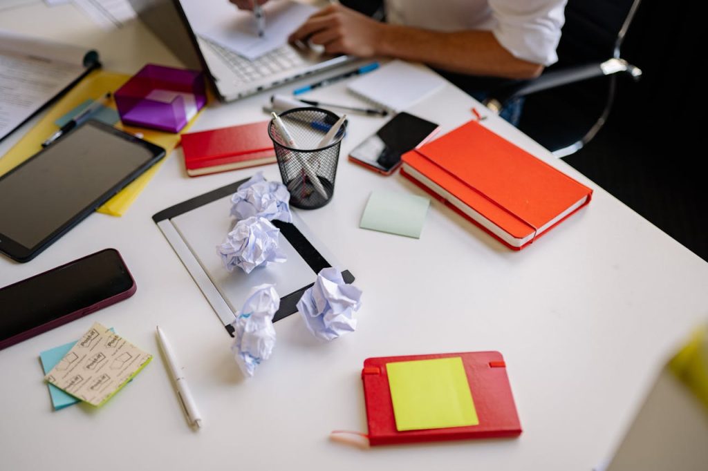 Messy white desk, scattered notebooks, crumpled papers, sticky notes, pens, a tablet, and a person working, indicating disorganization and clutter buildup