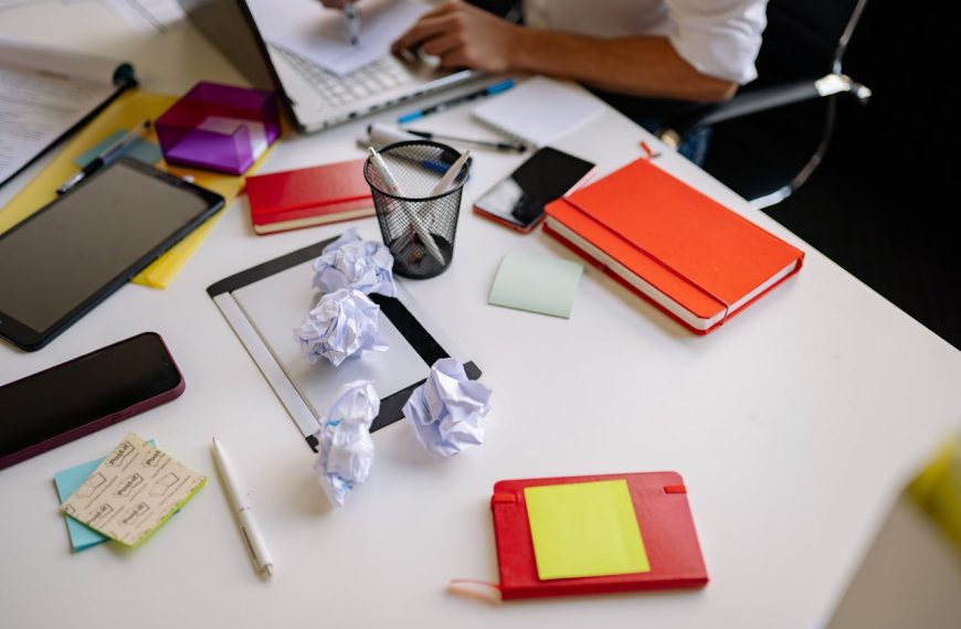 Messy white desk, scattered notebooks, crumpled papers, sticky notes, pens, a tablet, and a person working, indicating disorganization and clutter buildup