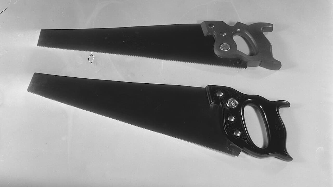 Two vintage hand saws, laid side by side on a flat surface, dark steel blades, wooden handles with finger holes, classic crosscut design, black and white image, carpentry tools