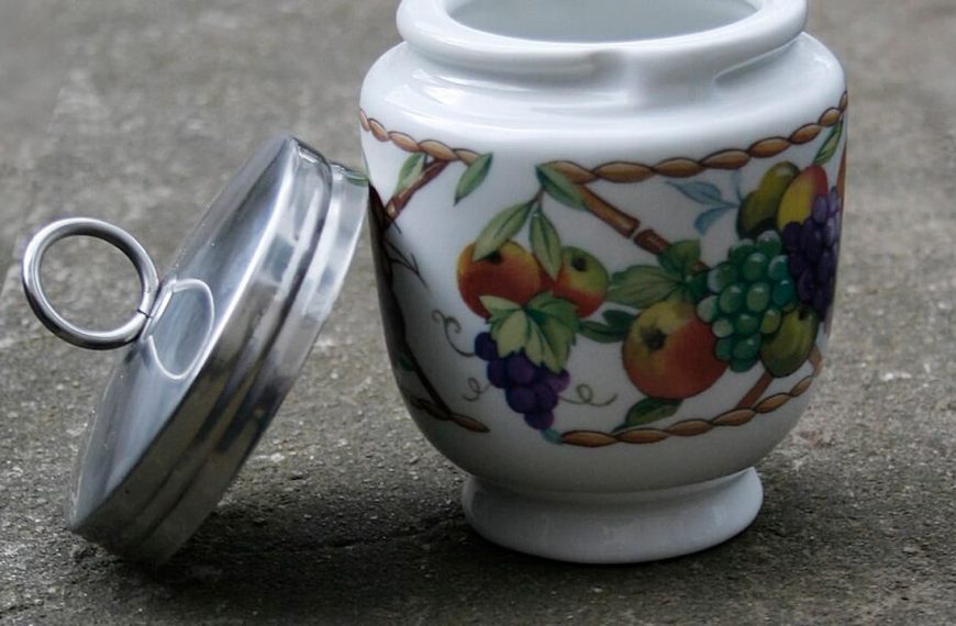 White ceramic jar with colorful fruit design, silver metal lid placed beside it, jar sitting on a concrete surface, partially open container