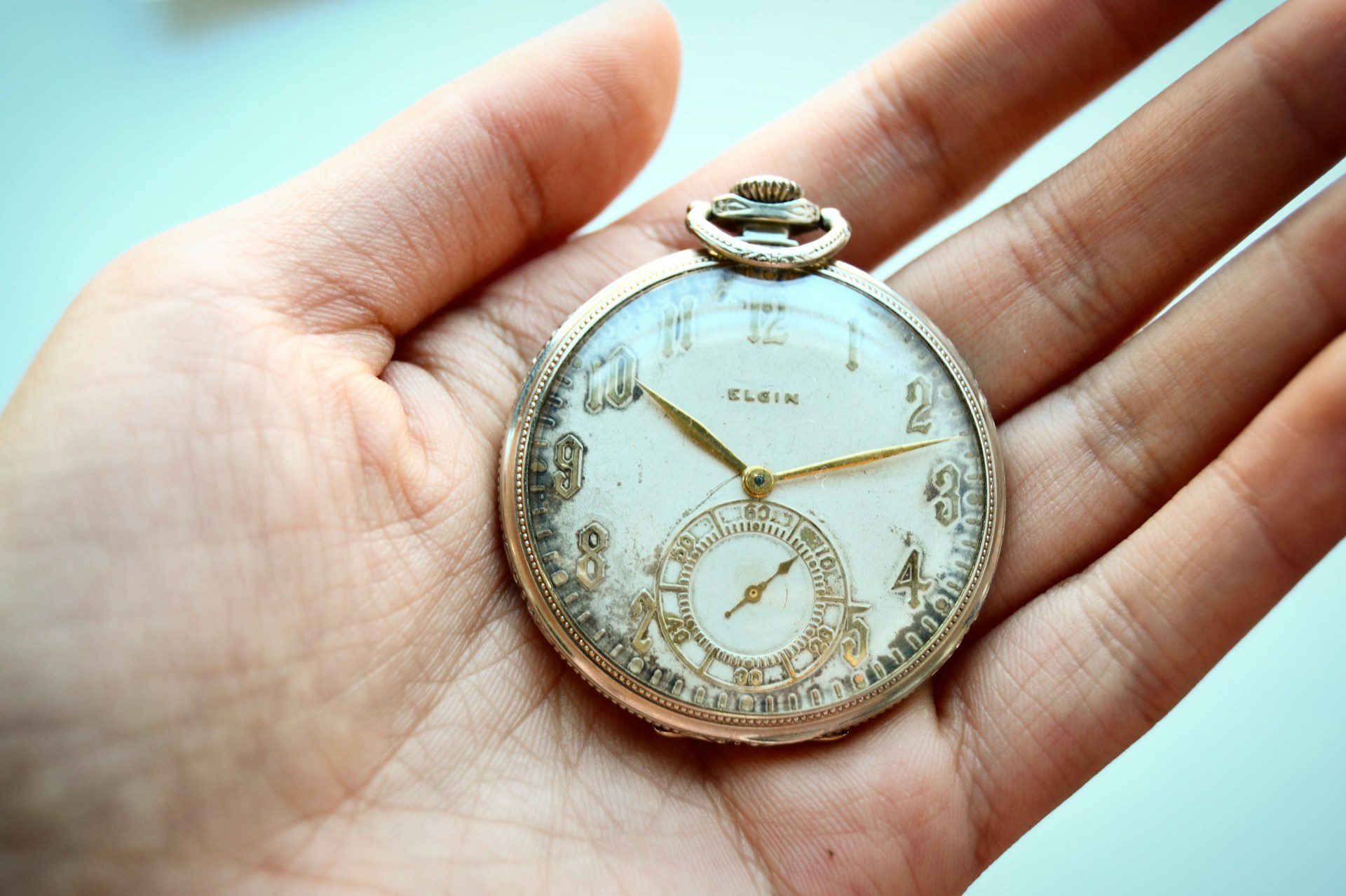 Close-up of a vintage pocket watch, gold detailing, white face with Roman numerals, ornate floral engravings, held gently in a person's palm, soft lighting, light blue background