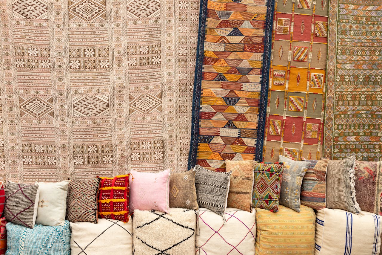 Display of handwoven textiles featuring large decorative rugs hanging on walls above rows of colorful cushions and pillows. Traditional geometric patterns in earth tones, oranges, and neutrals showcase artisanal craftsmanship