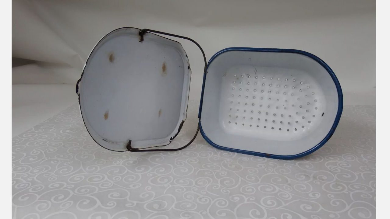 Pair of vintage white enamelware sink strainers with blue trim and rusted metal handles, displayed on a patterned surface