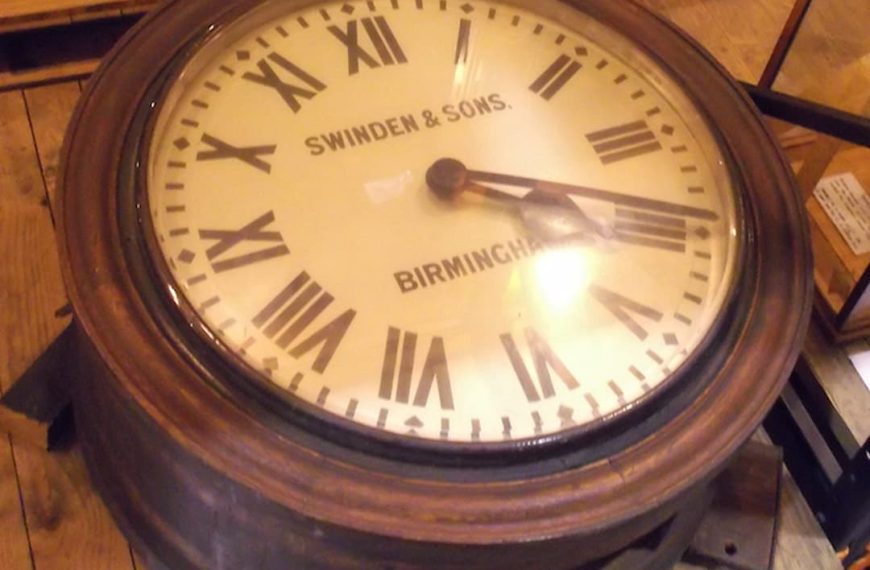 Large antique English dial clock, circular face with Roman numerals, labeled “Swinden & Sons, Birmingham,” set in a thick wooden frame, displayed on a wooden floor with supporting blocks and museum signage nearby