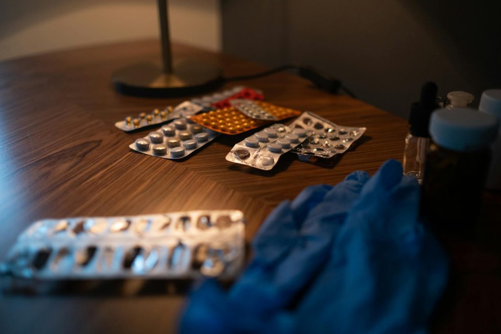 Assorted blister packs of medication and a pair of blue medical gloves on a wooden table