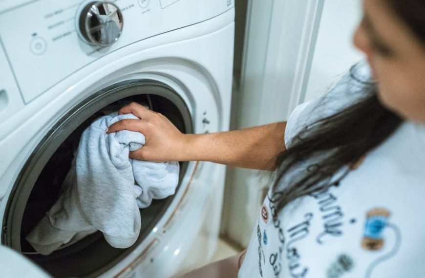 A woman loading light-colored clothing into a front-loading washing machine, suggesting the beginning of a laundry cycle