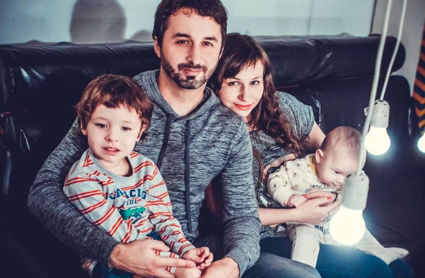 Smiling family of four sitting on a black couch, with the father holding a young boy and the mother holding a baby, surrounded by hanging light bulbs