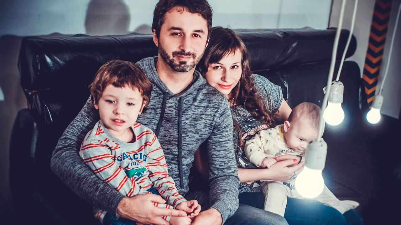 Smiling family of four sitting on a black couch, with the father holding a young boy and the mother holding a baby, surrounded by hanging light bulbs