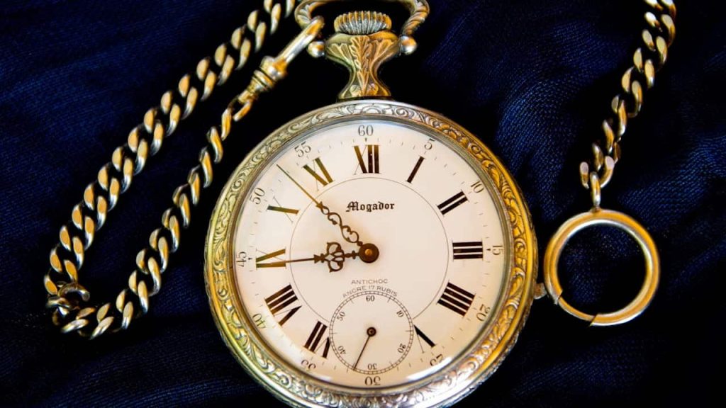 A vintage gold pocket watch with Roman numerals and a decorative chain, lying on a dark fabric background