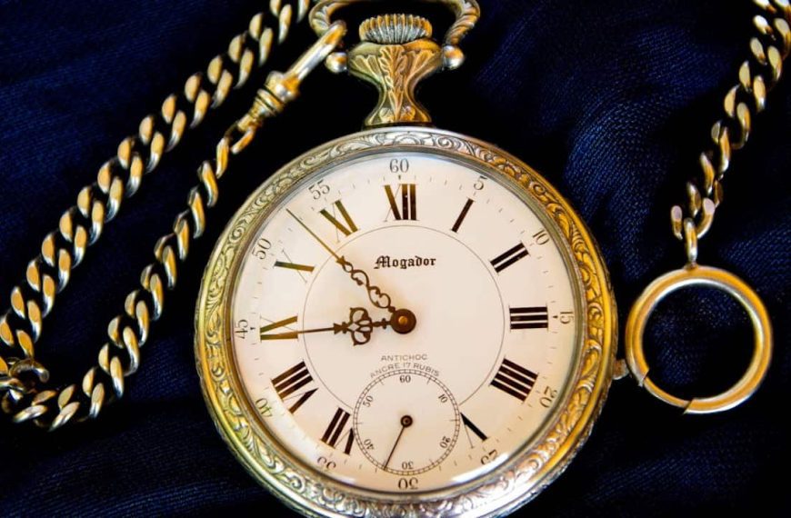 A vintage gold pocket watch with Roman numerals and a decorative chain, lying on a dark fabric background