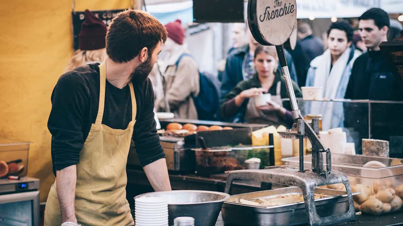 Street food vendor in a yellow apron stands behind a counter with a potato cutter, food containers, and paper cups, while customers queue in front of the stall labeled "The Frenchie