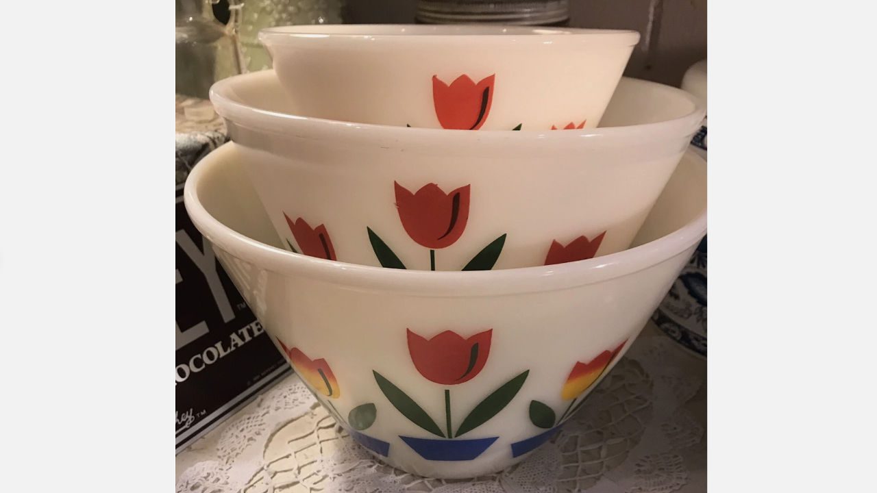 Stack of vintage Fire King mixing bowls with red tulip floral designs on a white glass base, resting on a lace tablecloth