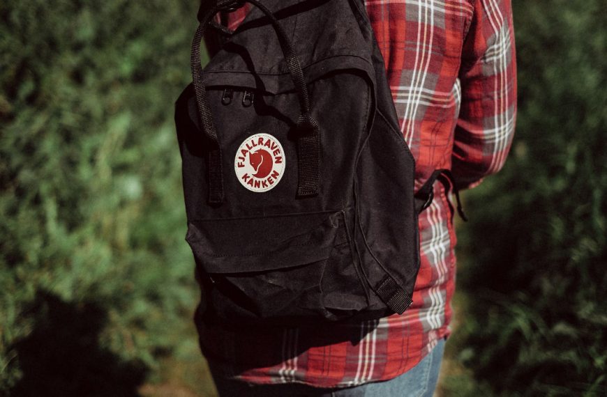 Black Fjällräven Kånken backpack worn by a person in a red plaid shirt, walking outdoors