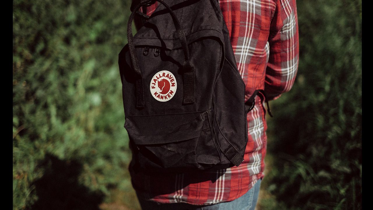 Black Fjällräven Kånken backpack worn by a person in a red plaid shirt, walking outdoors