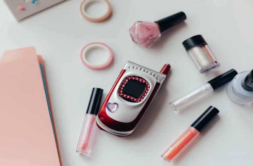 Decorated red and silver flip phone, surrounded by lip gloss tubes, nail polish bottles, pink file folder, and rolls of decorative tape, on a white surface