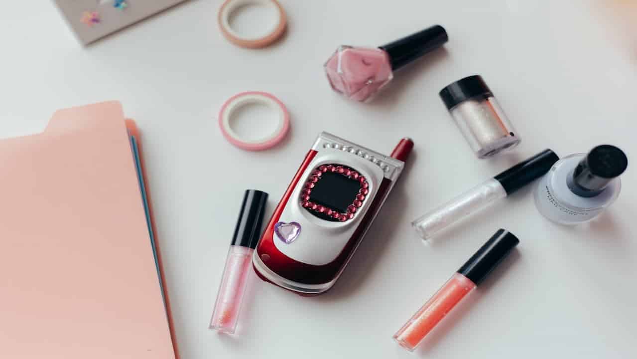 Decorated red and silver flip phone, surrounded by lip gloss tubes, nail polish bottles, pink file folder, and rolls of decorative tape, on a white surface