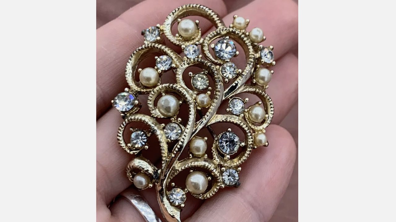 Vintage floral brooch with intricate gold-tone swirls, adorned with faux pearls and round clear rhinestones, held in hand against a natural background