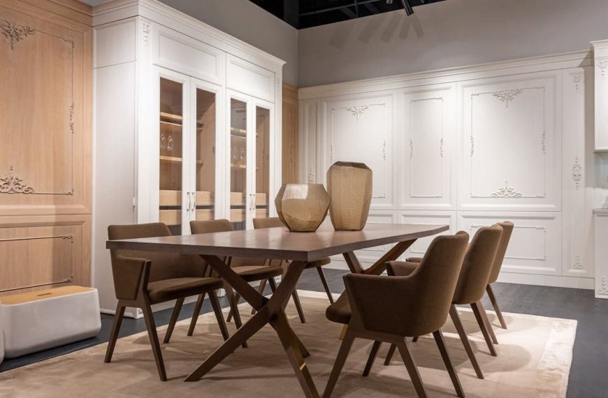 Dining room with a wooden table, six upholstered chairs with angled legs, two decorative vases on the table, large built-in white cabinetry with glass doors and intricate molding, soft beige rug underneath, warm ambient lighting, neutral color palette with wood and white tone